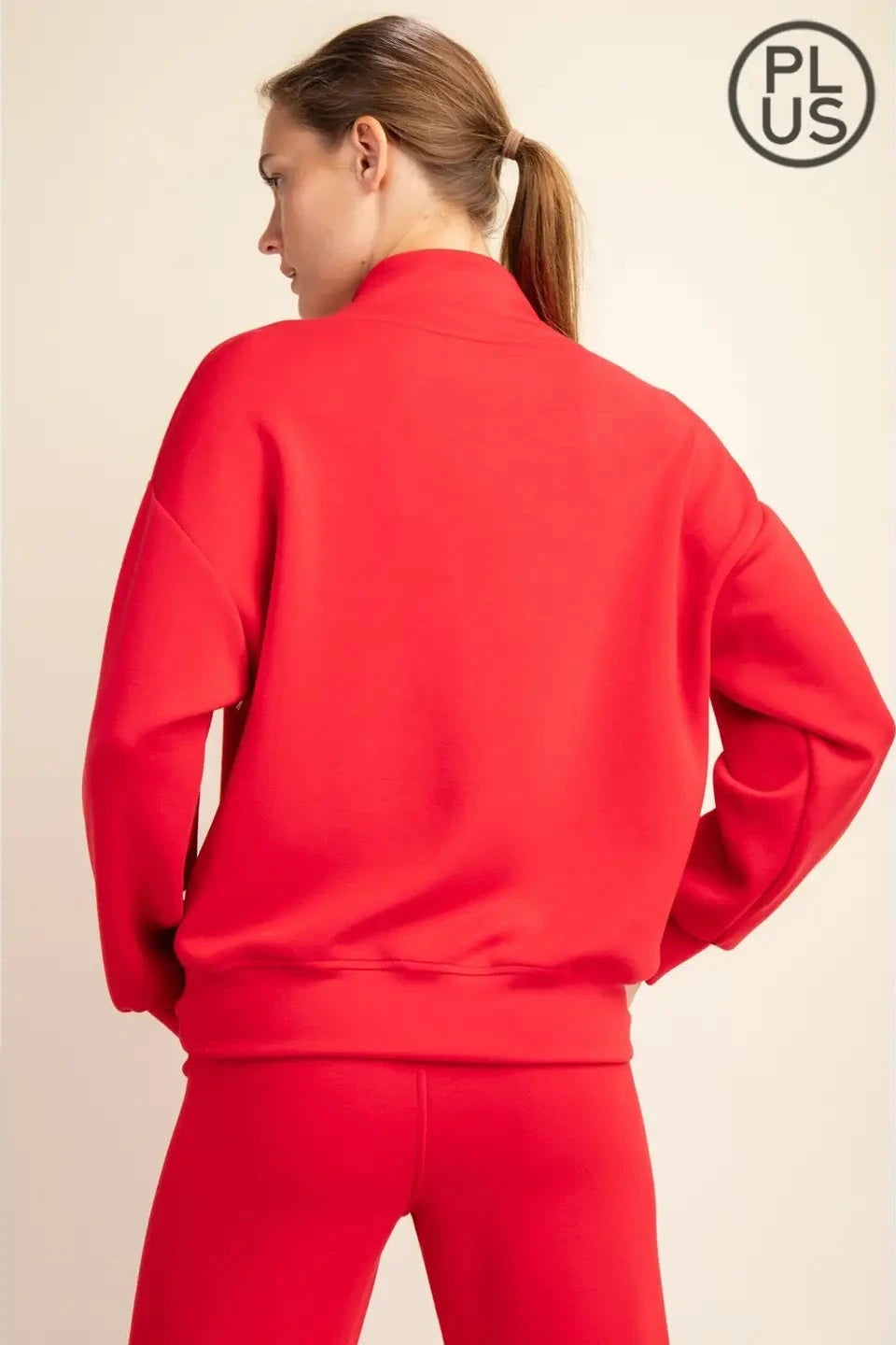 A woman shown from the back wearing a bright red sweatshirt and matching red pants against a light beige studio background. A circular 'PLUS' logo appears in the top right corner.