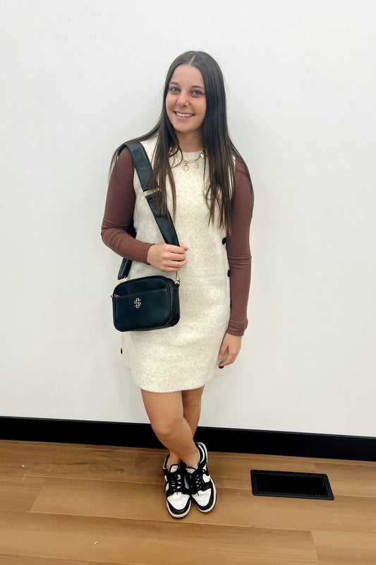 Woman in a white dress with a black handbag standing against a plain wall.