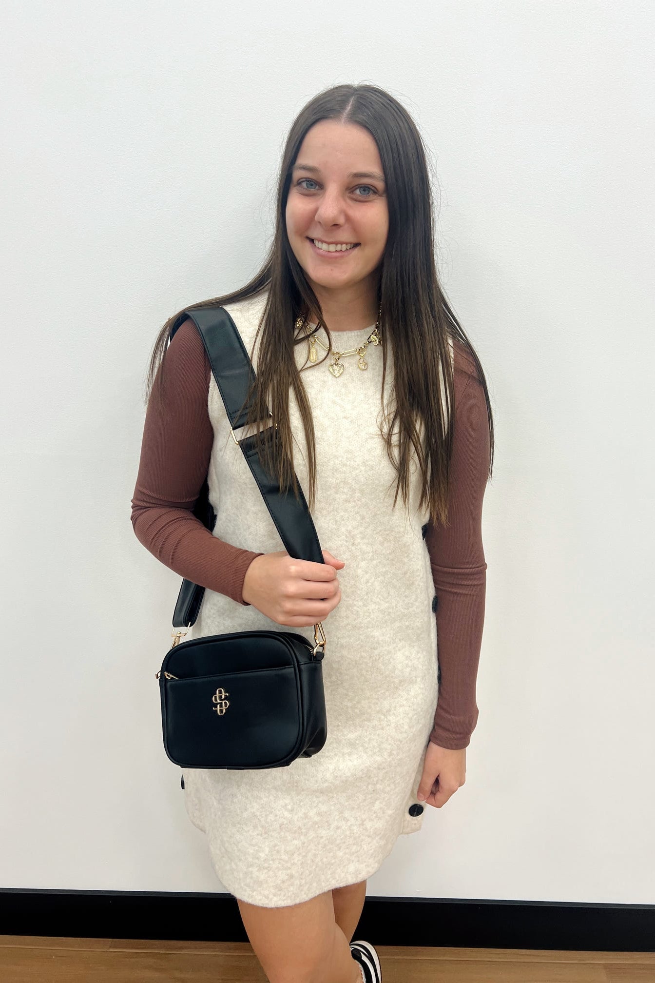 Woman in a white dress with brown long-sleeve underneath, holding a black handbag against a plain background