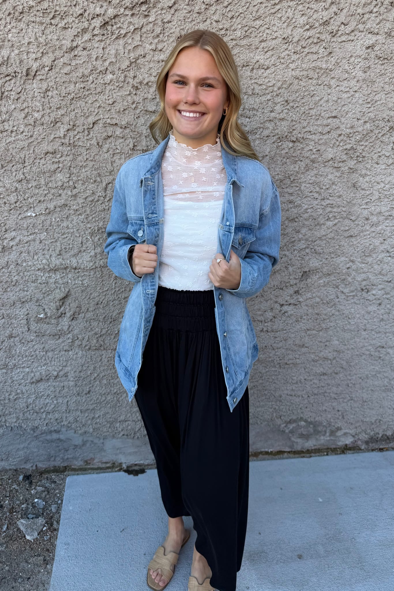 Woman wearing a denim jacket, white top, and black pants standing against a textured wall.