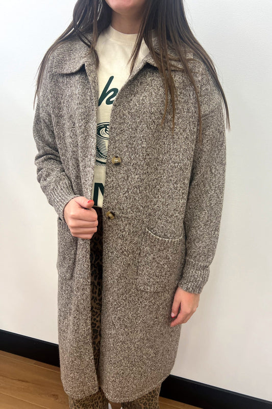 Person wearing a long brown coat over a graphic t-shirt against a white wall.