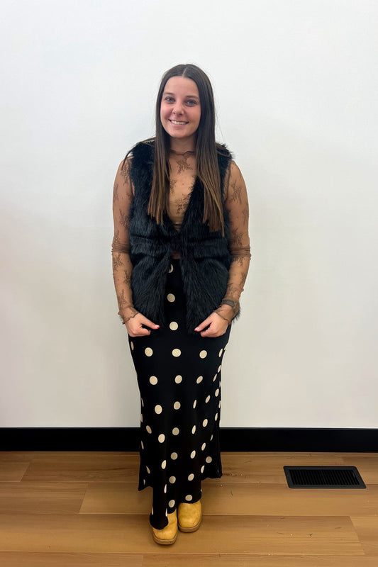 Person wearing a black fur vest over a polka dot dress on a plain background