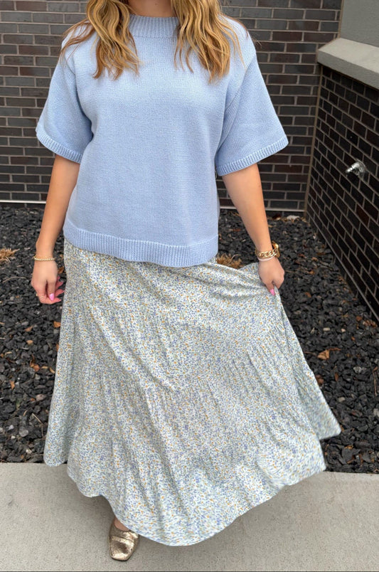 Woman wearing a light blue sweater and silver skirt standing against a brick wall.
