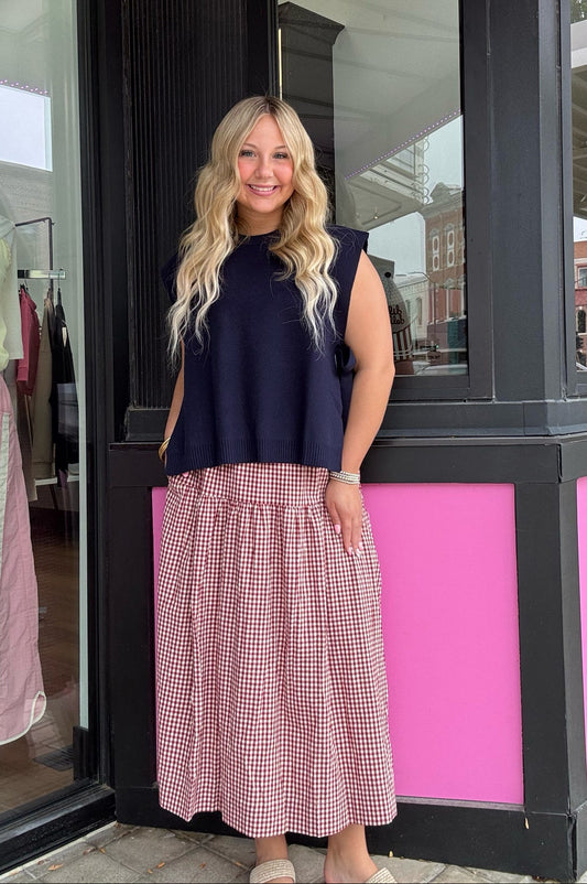 A woman stands in front of a storefront wearing a sleeveless navy top and a pink and white gingham midi skirt. The shop has black-framed windows and bright pink paneling.