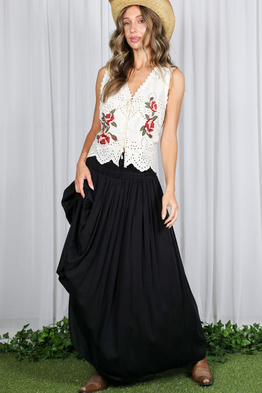 Woman wearing a white embroidered top and black skirt with a straw hat, standing against a white curtain.