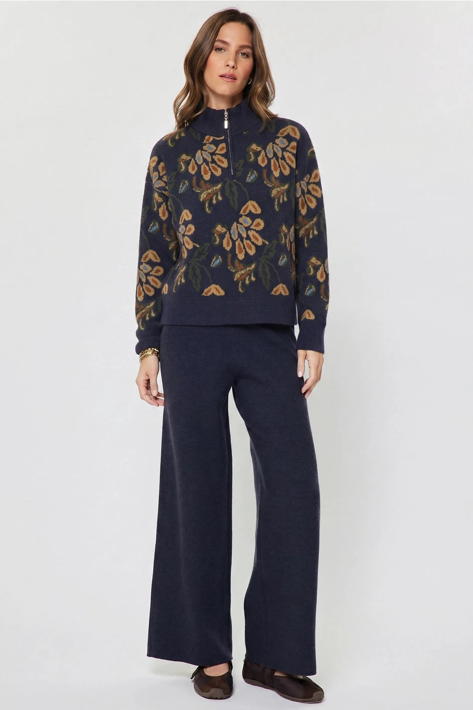 A woman modeling a navy blue knit half-zip sweater with a brown and gold floral pattern, paired with matching navy wide-leg trousers, against a light gray studio background.