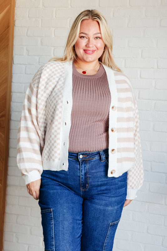Woman wearing a striped cardigan, brown sweater, and blue jeans against a white brick wall.