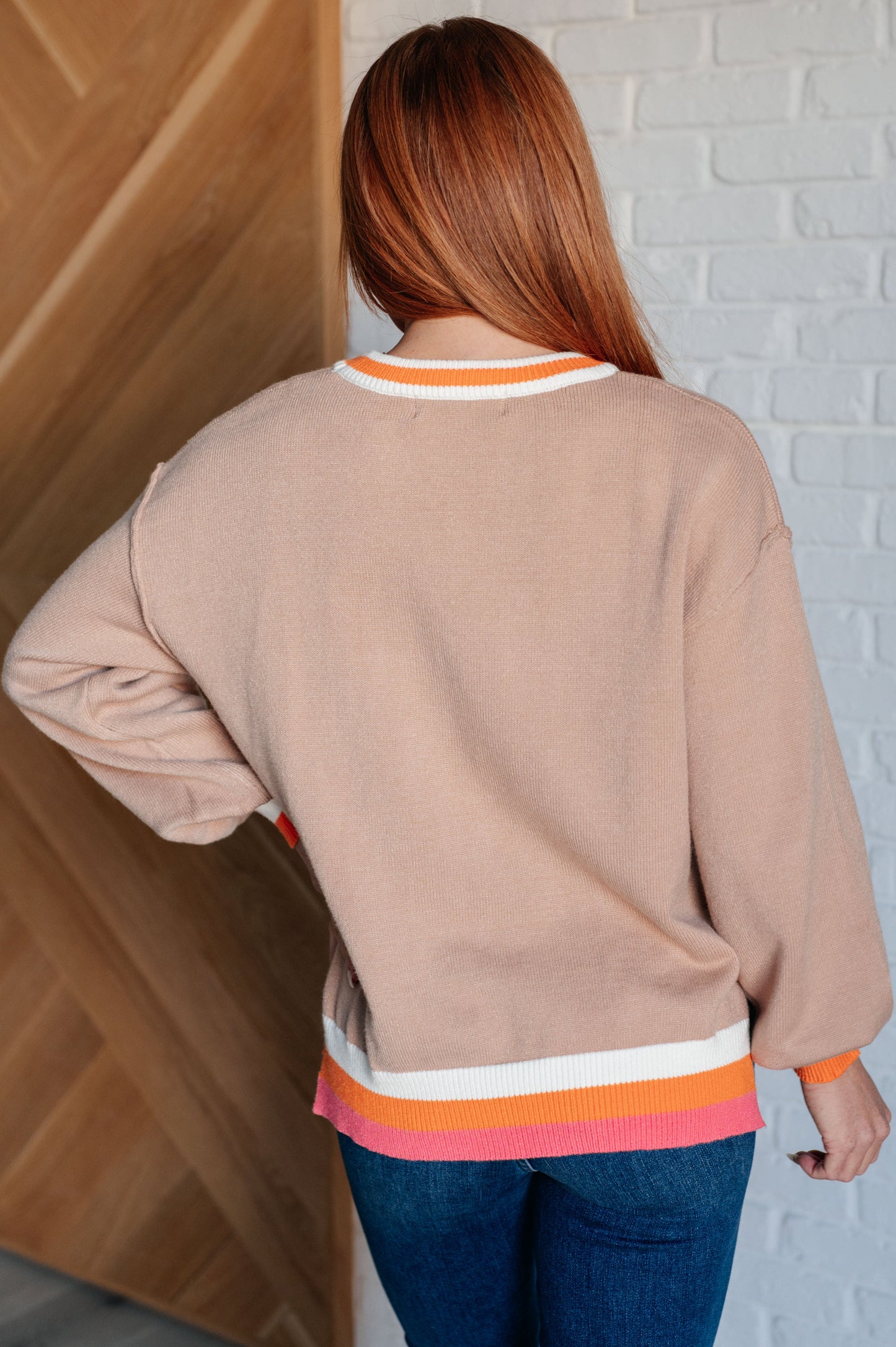 Woman wearing a beige sweater with colorful stripes, standing against a white brick wall.