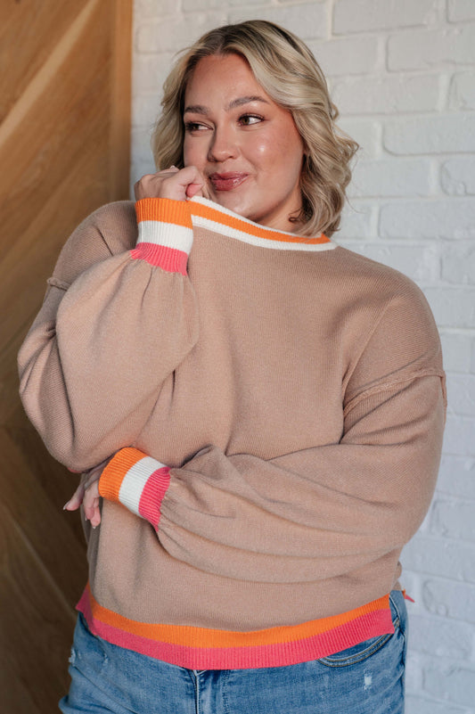 Woman wearing a beige sweater with colorful cuffs and hem against a white brick wall.