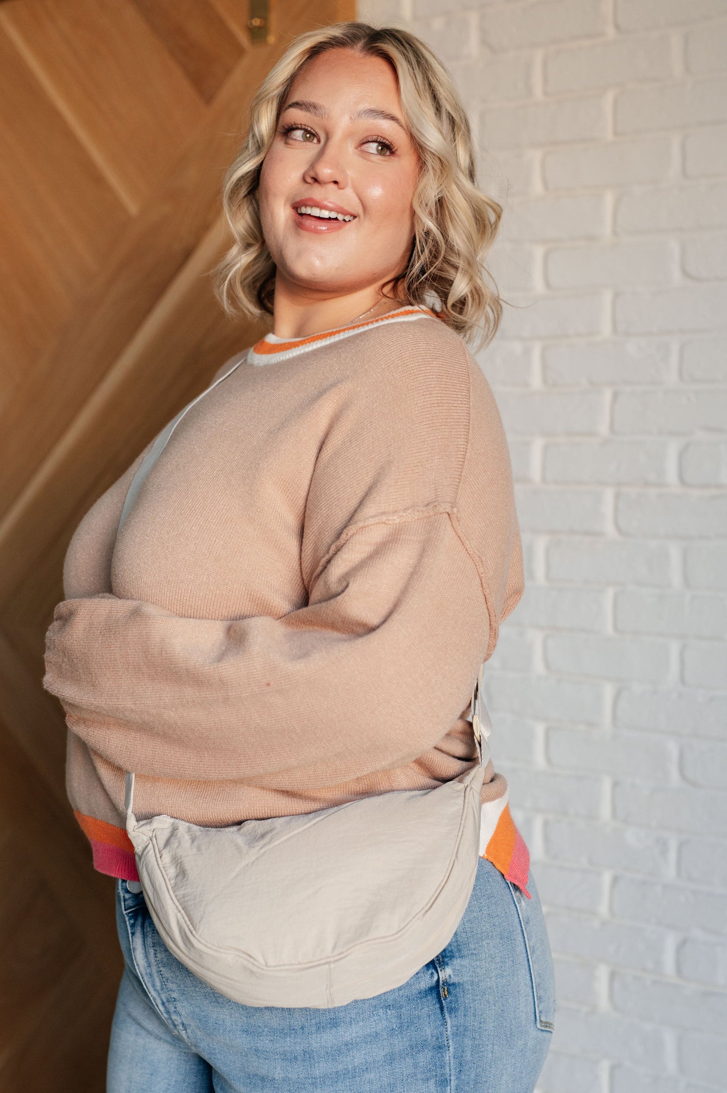 Woman wearing a beige sweater and blue jeans against a white brick wall.