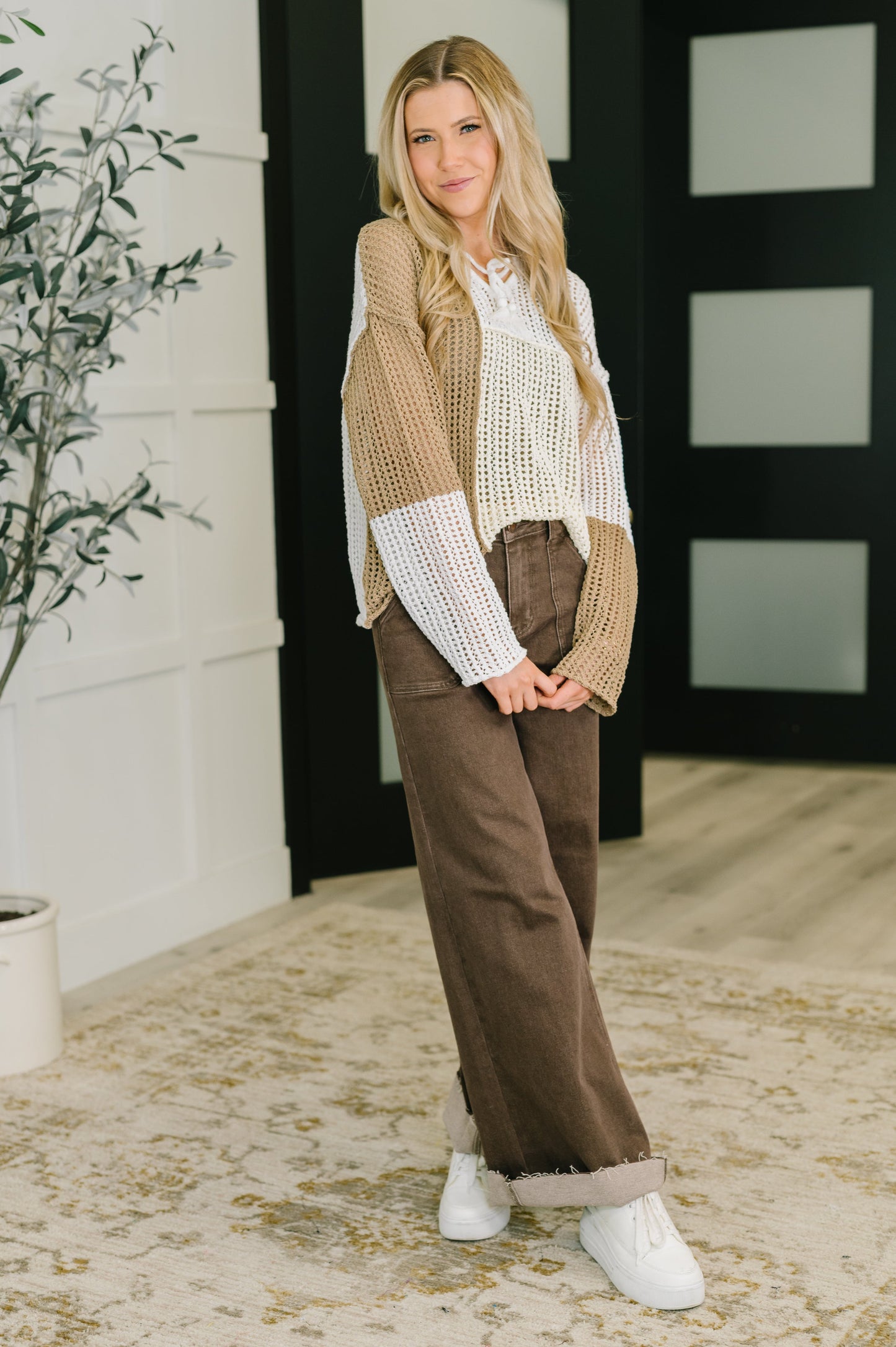 Woman wearing a beige and brown outfit standing in a room with a plant and door in the background.