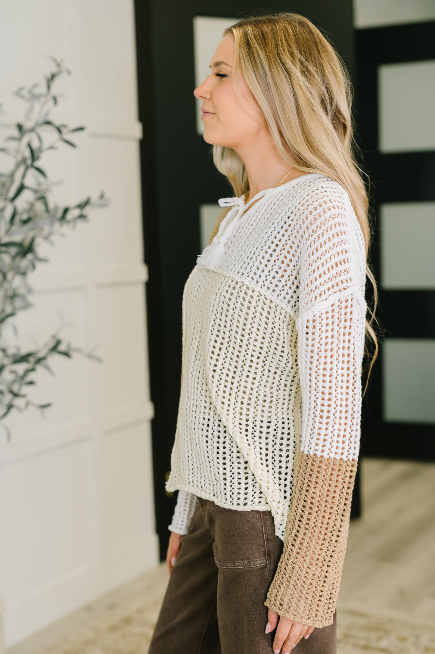 Woman wearing a white and beige crochet top with brown pants in a room.