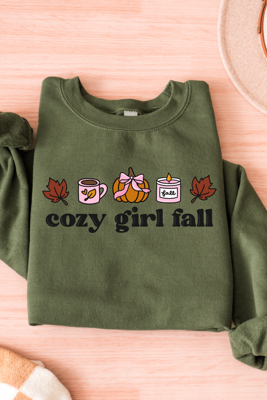 Green sweatshirt with 'cozy girl fall' text and autumn-themed graphics on a pink surface.