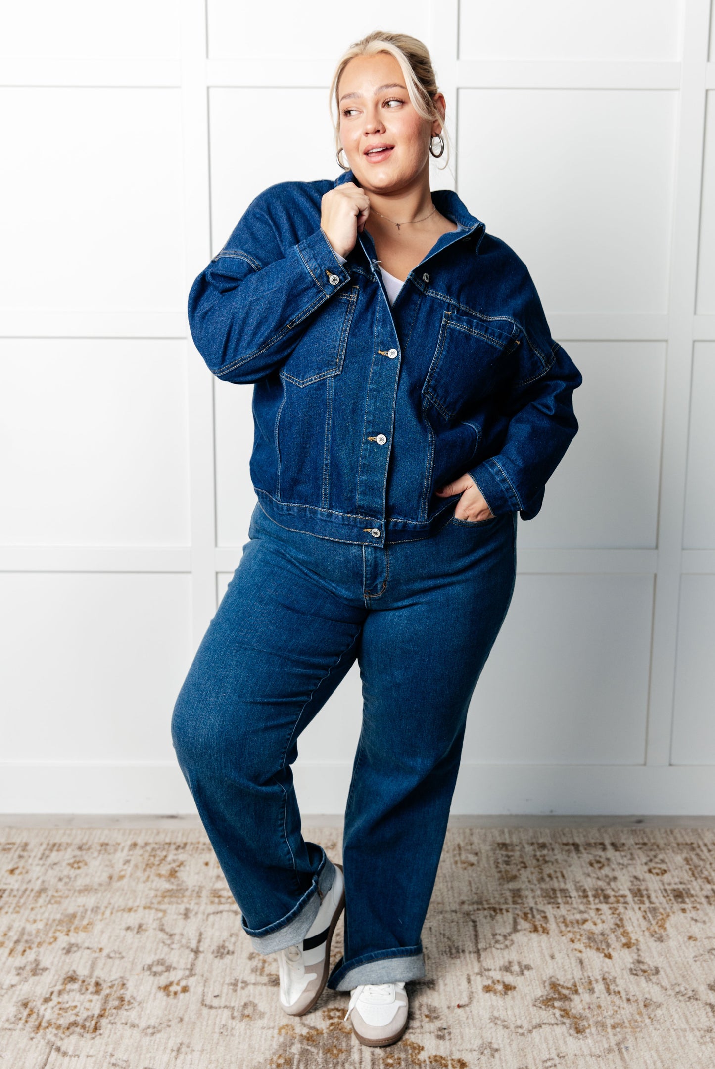 Person wearing a blue denim jacket and jeans against a white tiled wall.