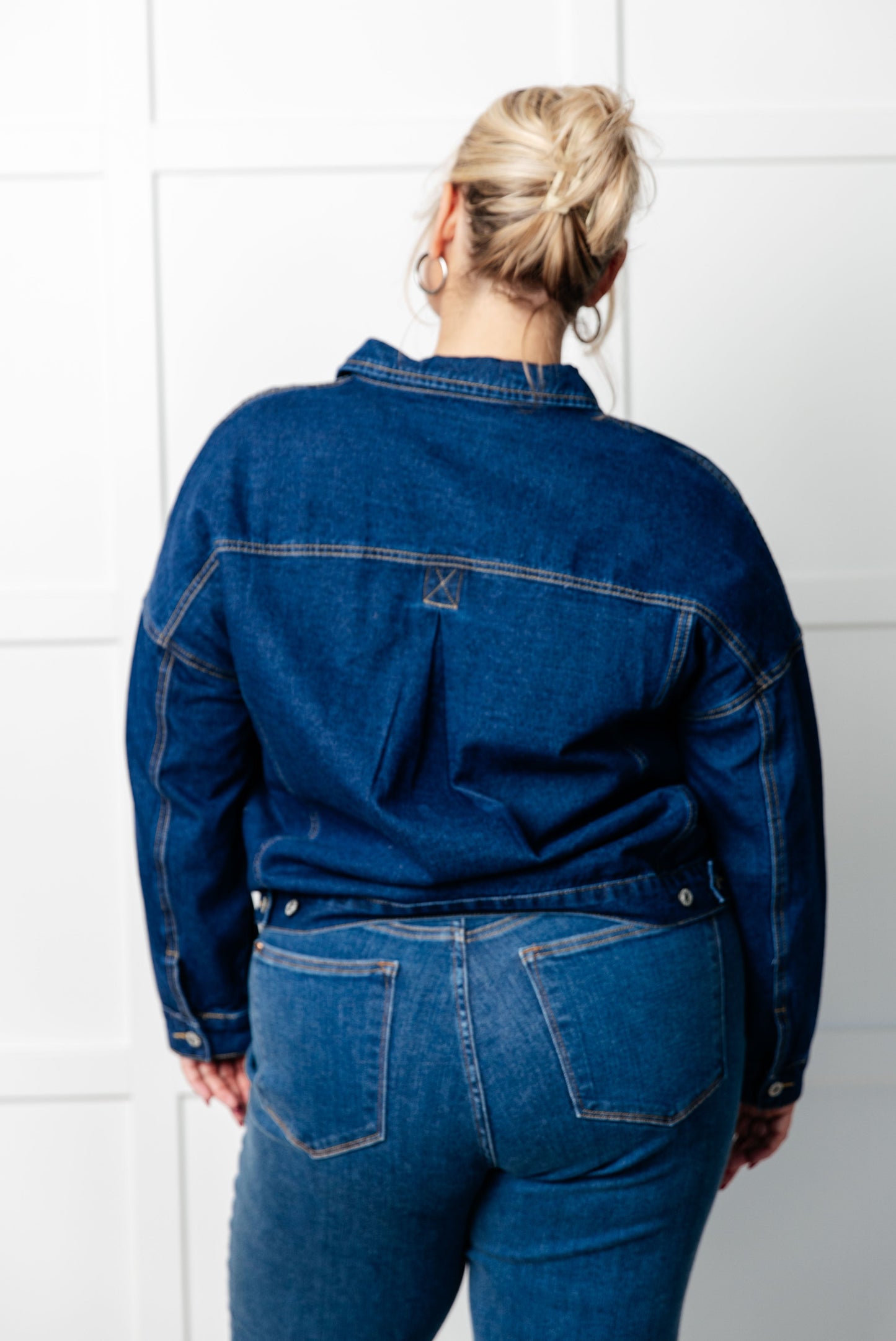 Person wearing a blue denim jacket and jeans against a white tiled wall.