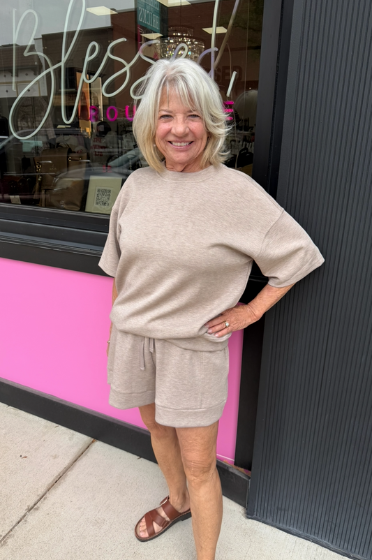 A woman with short gray hair stands outdoors in front of a storefront, wearing a matching beige lounge set consisting of an oversized short-sleeve top and shorts. The storefront has a bright pink lower section and a dark gray vertical panel on the right.