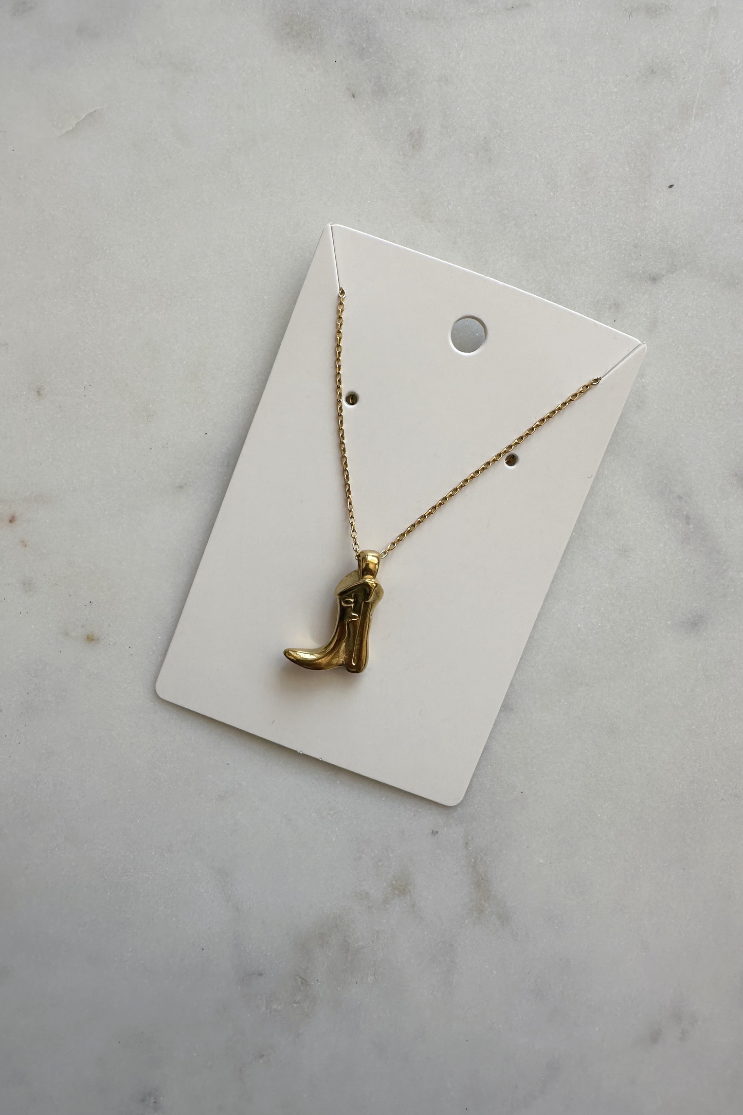 A golden cowboy boot necklace with an adjustable stainless steel chain, displayed on a white card against a marble background.