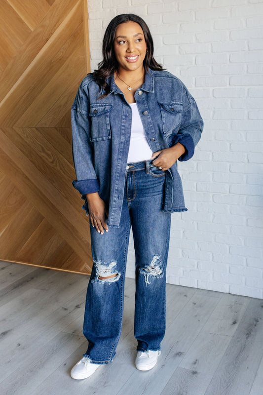 Woman wearing a denim jacket and jeans standing against a white brick wall.