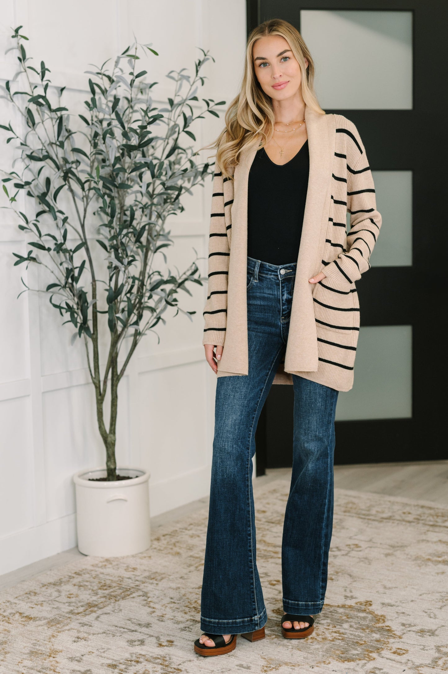 Woman wearing a beige cardigan with black stripes, black top, and blue jeans standing indoors.