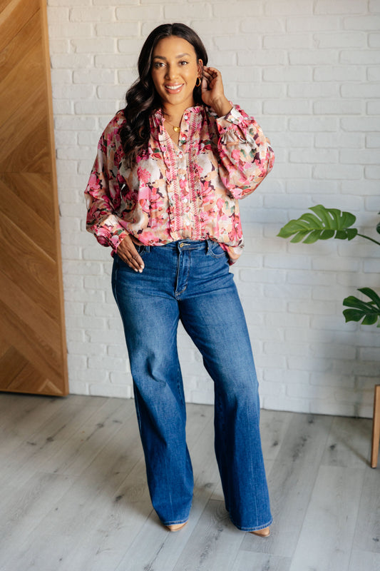 Woman wearing a floral blouse and blue jeans standing indoors against a white brick wall.