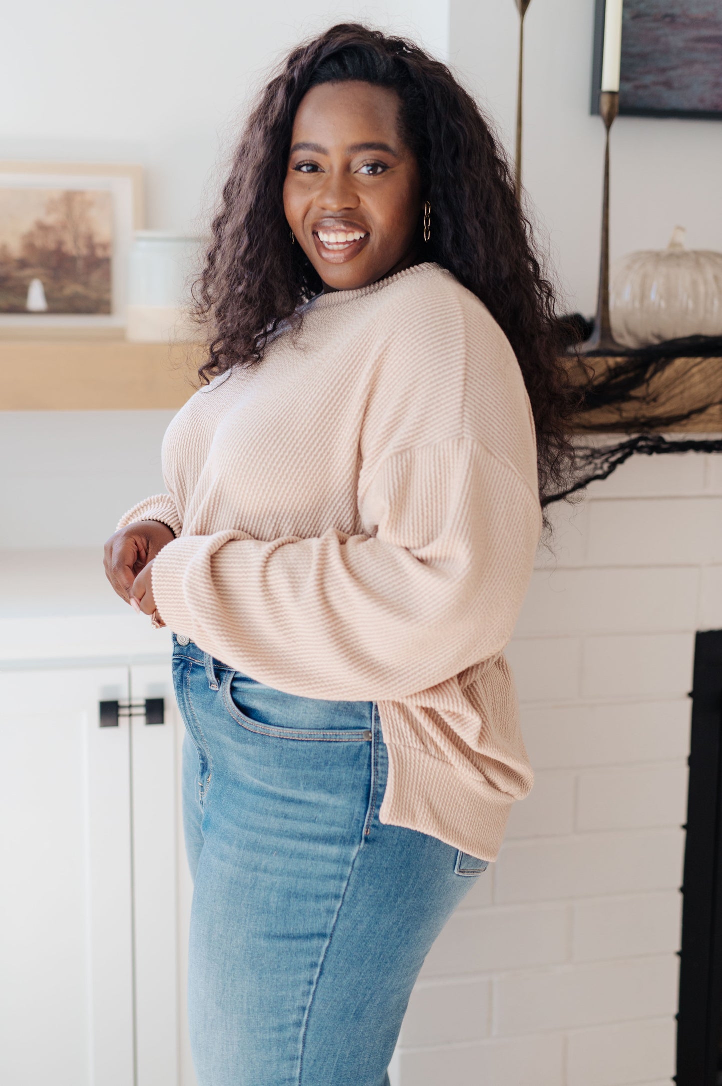 Woman wearing a beige sweater and blue jeans standing in a room with a fireplace.