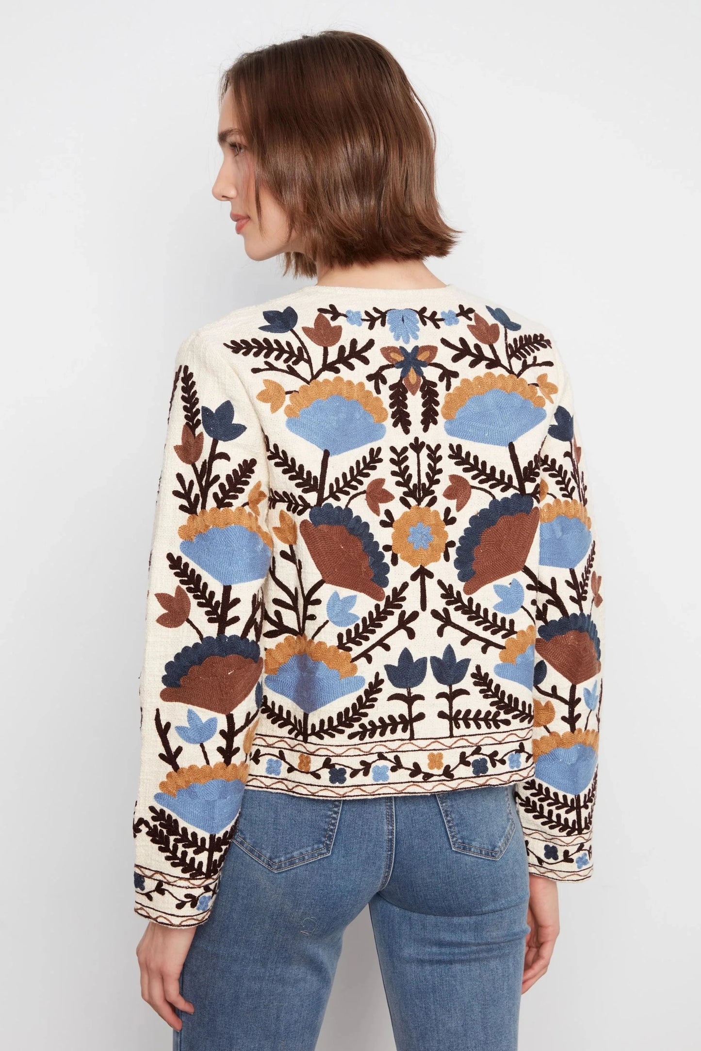 A woman shown from the back wearing a cream-colored cropped sweater with a colorful embroidered bohemian pattern in blue, brown, and orange, paired with blue jeans against a plain white background.