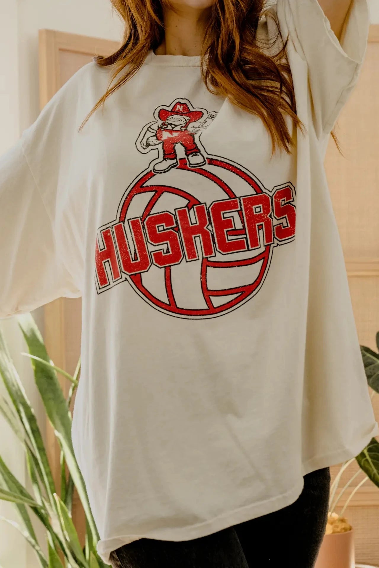 A woman wearing an oversized cream-colored t-shirt with a red and black 'HUSKERS' graphic featuring a mascot character on a volleyball, set against a warm indoor background with plants.