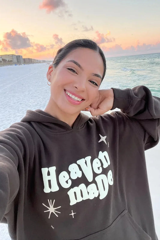 Person wearing a hoodie with 'Heaven made' text at the beach during sunset.