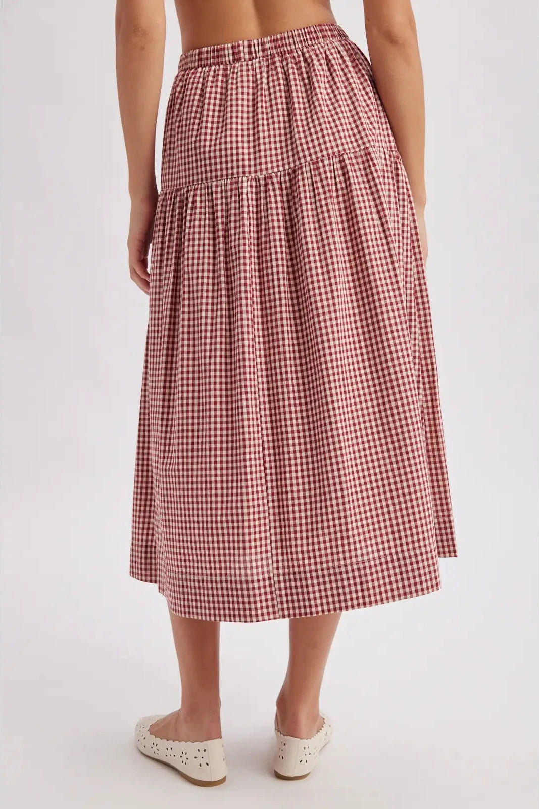 A model from the back wearing a tiered midi skirt with a red and white gingham pattern, paired with white flat shoes, against a light gray studio background.