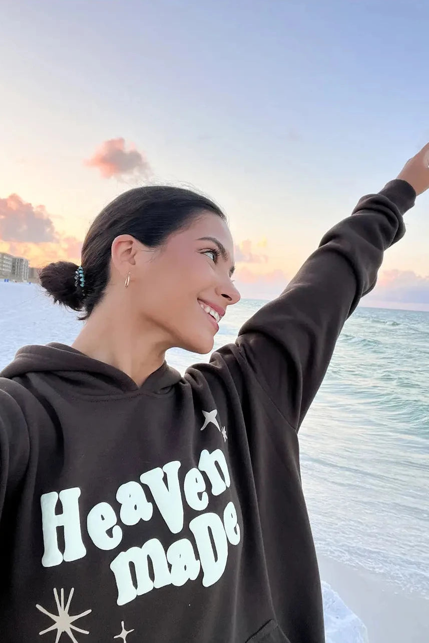 Woman wearing a hoodie with 'Heaven Made' text by the ocean at sunset
