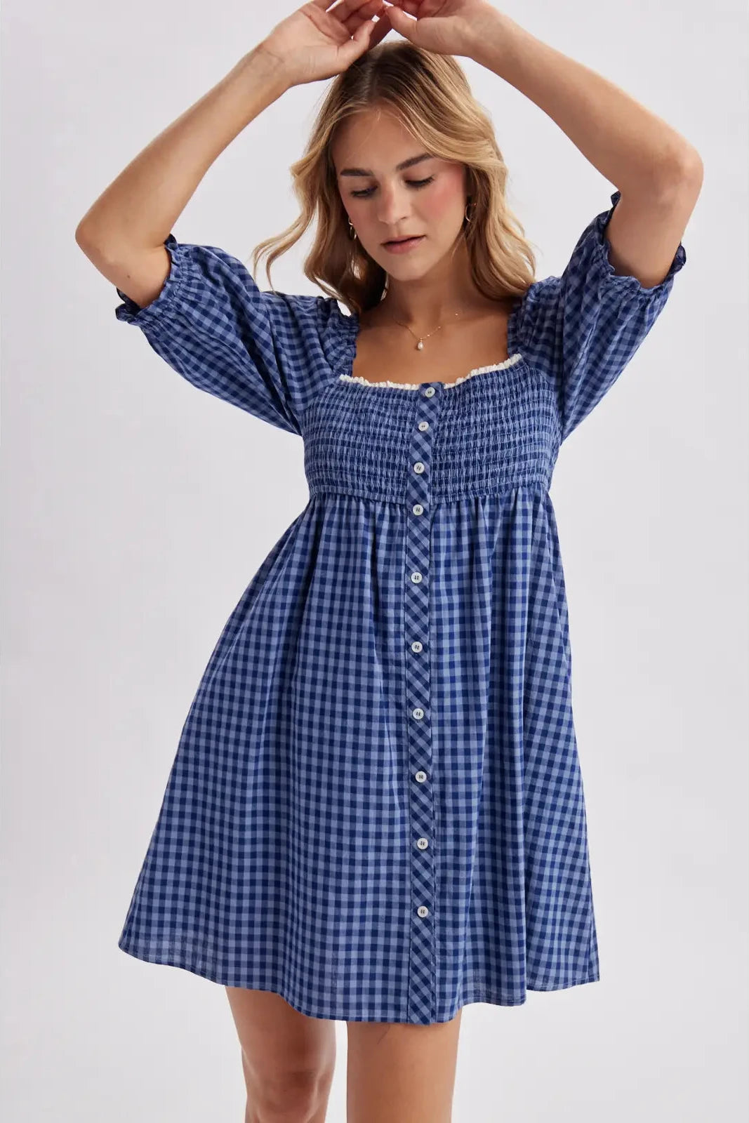 A woman modeling a blue and white gingham mini dress with puff sleeves, a smocked bodice, and a button-down front, set against a plain light gray studio background.