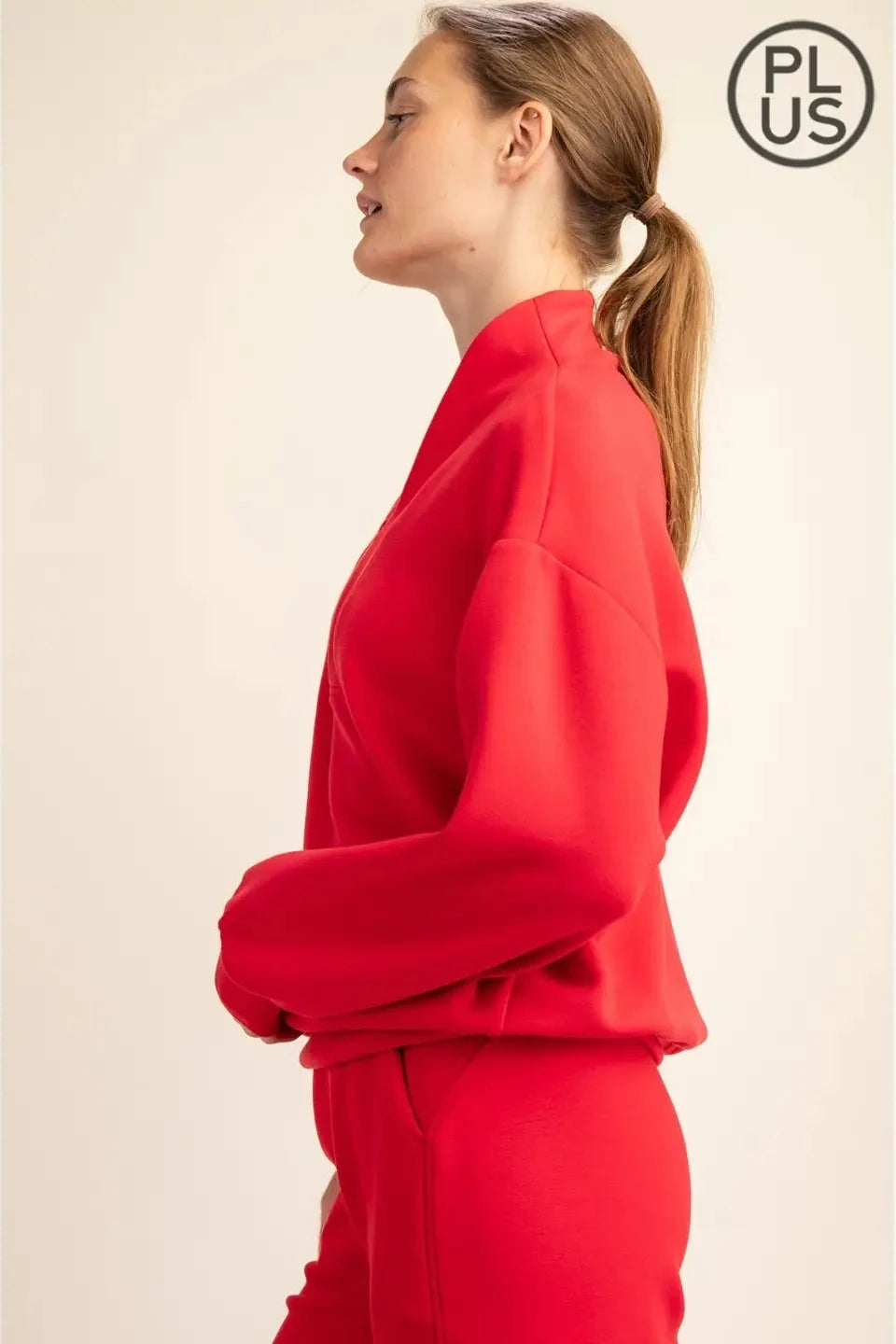 A woman in side profile wearing a bright red knit top and matching red bottoms, photographed against a plain light beige studio background. A circular 'PLUS' mark appears in the top-right corner.