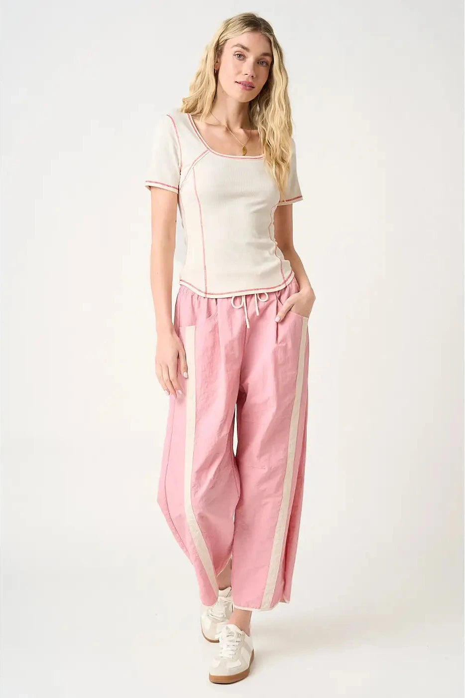 A woman modeling a two-piece loungewear set consisting of a cream short-sleeve top with pink piping and matching pink wide-leg pants with cream side stripes, paired with white lace-up shoes, against a plain light background.