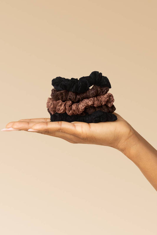 A hand holding a small stack of four velvet hair scrunchies in black and brown shades, set against a solid beige background.