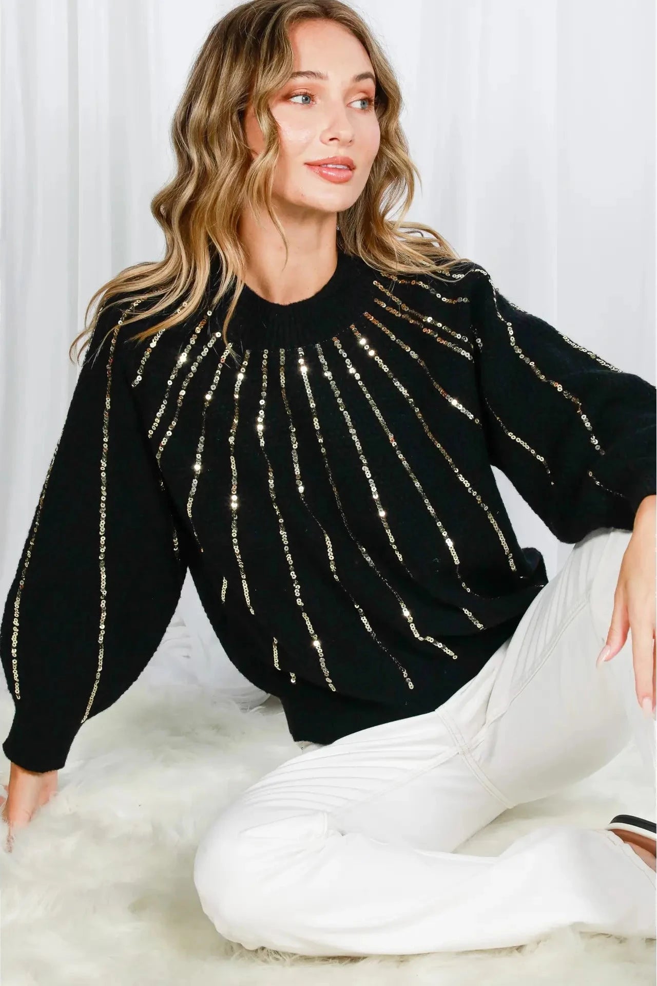 Woman wearing a black sweater with gold stripes sitting on a white surface.