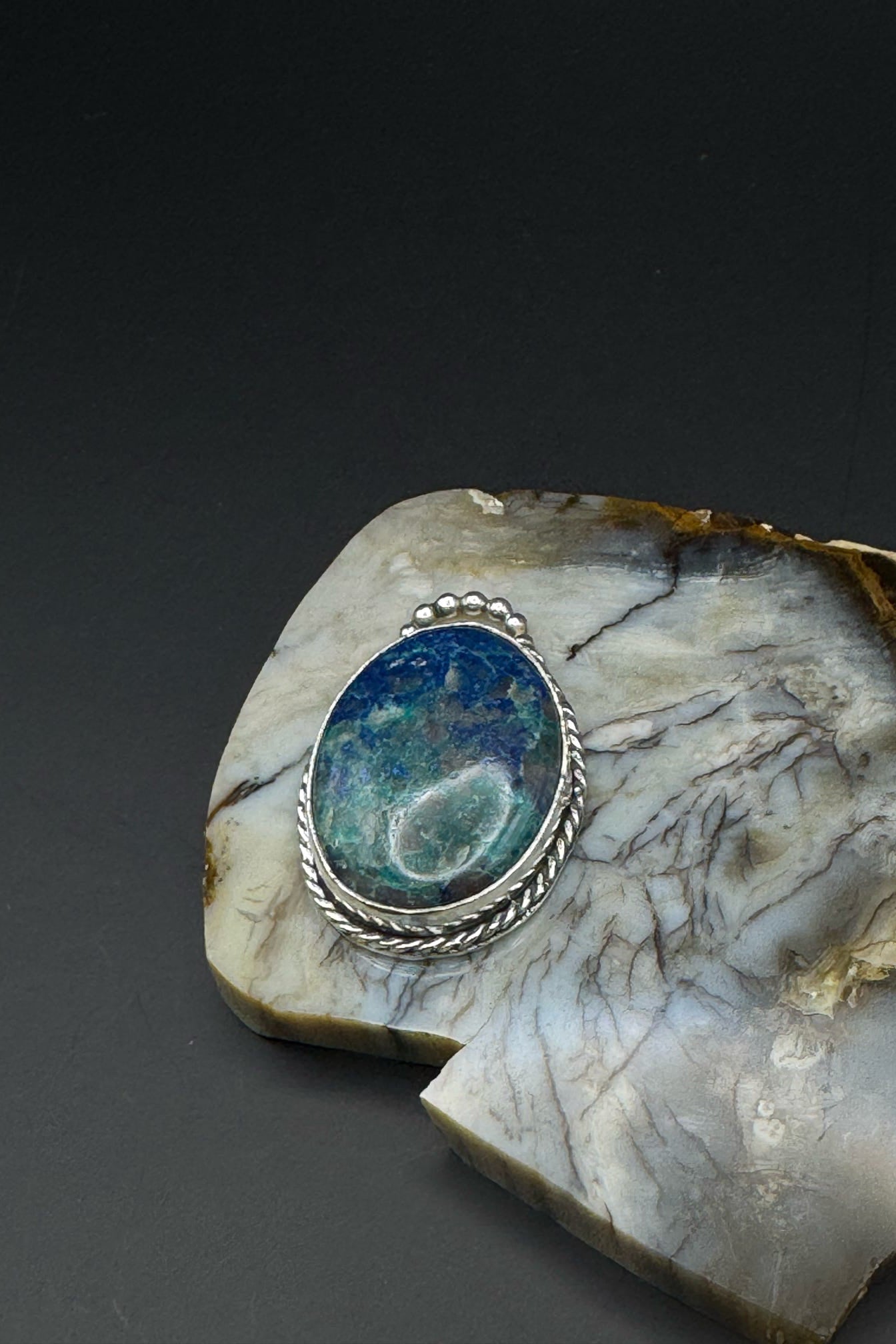 Stylized image of a gemstone in a silver setting on a textured stone surface with a dark background.