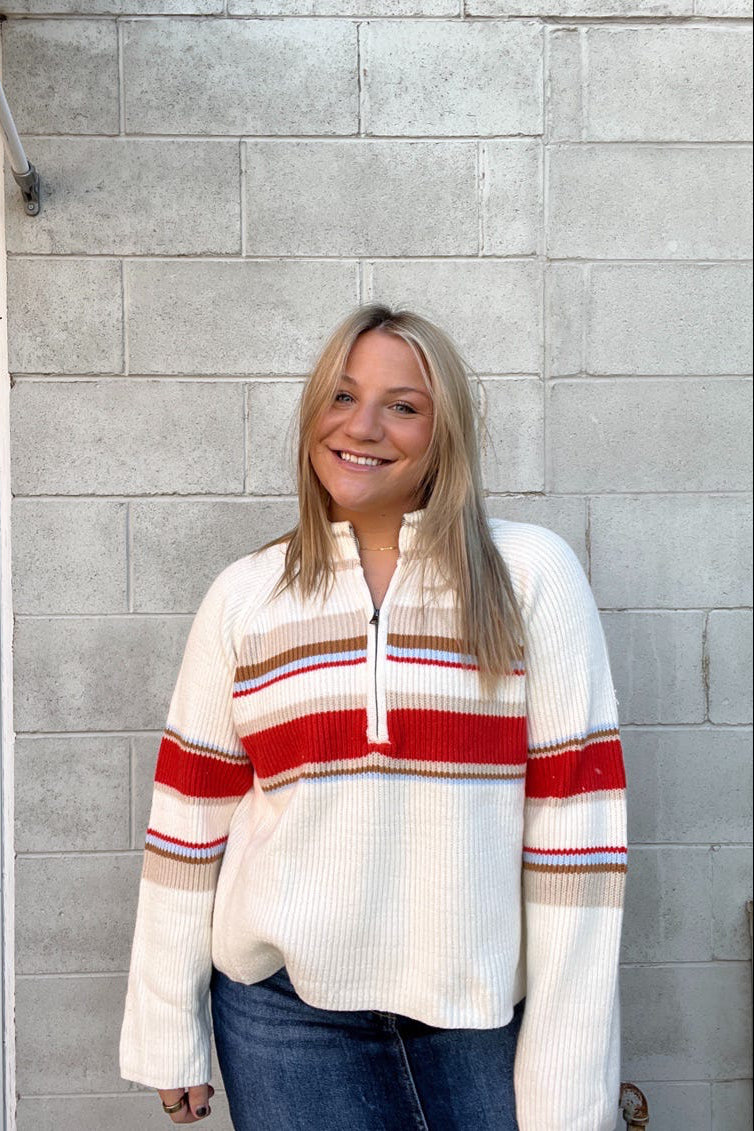 Woman wearing a white sweater with red and brown stripes against a gray brick wall.