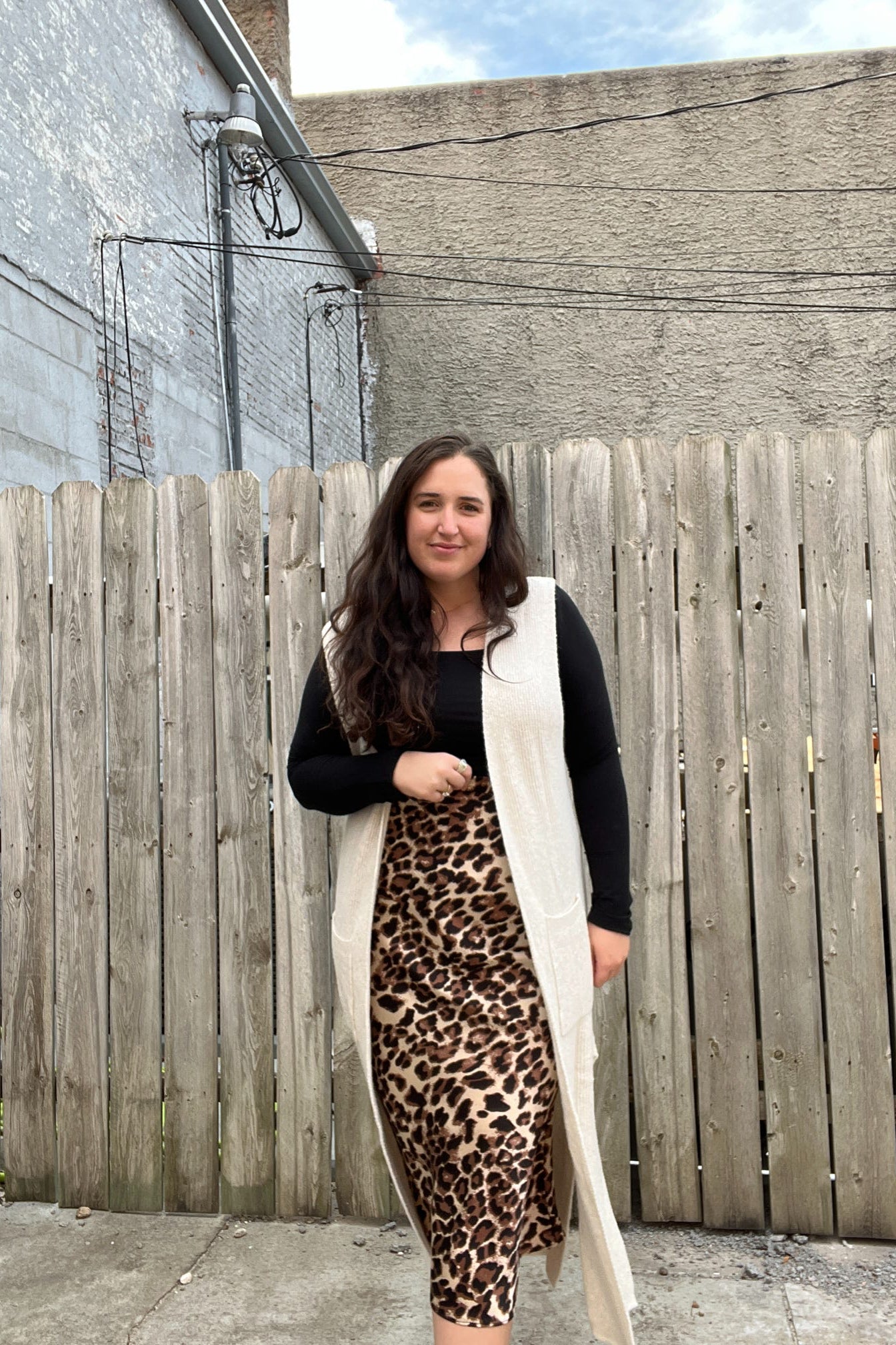 Woman wearing a long cream scarf, black top, and leopard print skirt standing in front of a wooden fence.