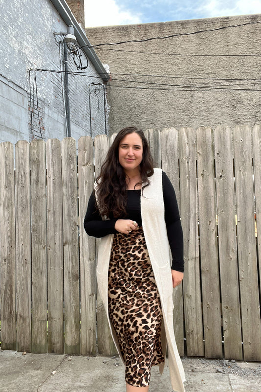 Woman wearing a long cream scarf, black top, and leopard print skirt standing in front of a wooden fence.