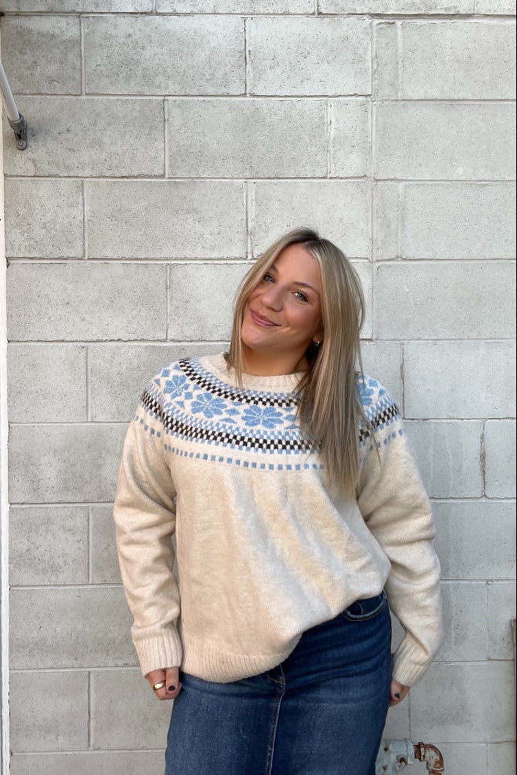 Woman wearing a cream sweater with blue and black patterns against a gray brick wall.
