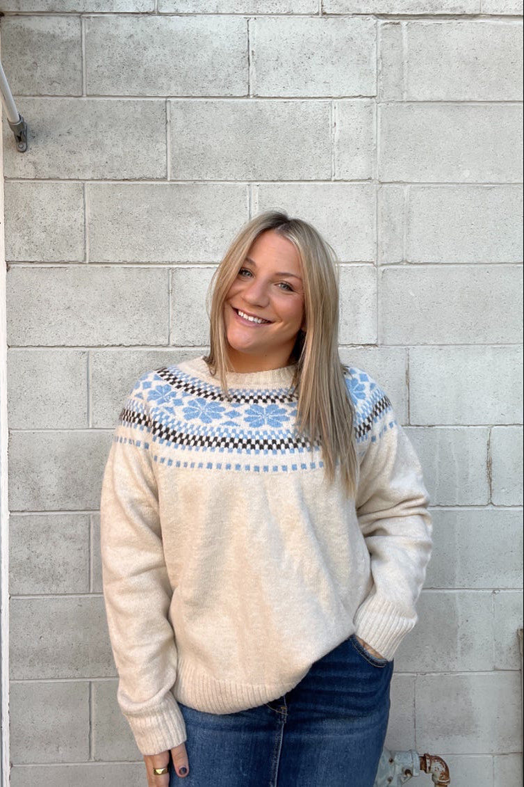 Woman wearing a cream sweater with blue patterns against a gray brick wall.