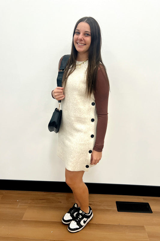 Woman wearing a cream dress with black buttons and brown sleeves, holding a black bag indoors.