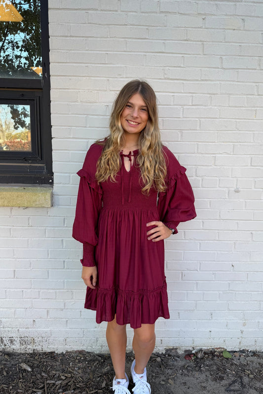 Woman wearing a burgundy dress standing against a white brick wall.