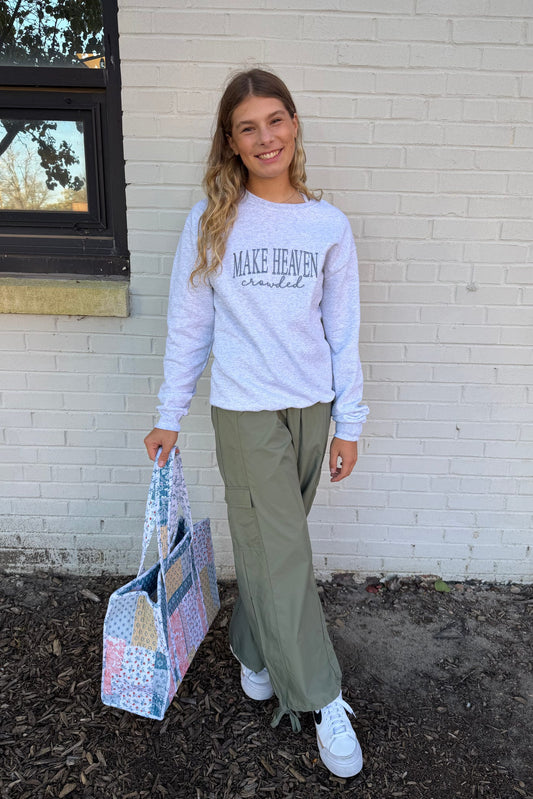 Person wearing a light gray sweatshirt with text, holding a patterned bag, standing against a white brick wall.