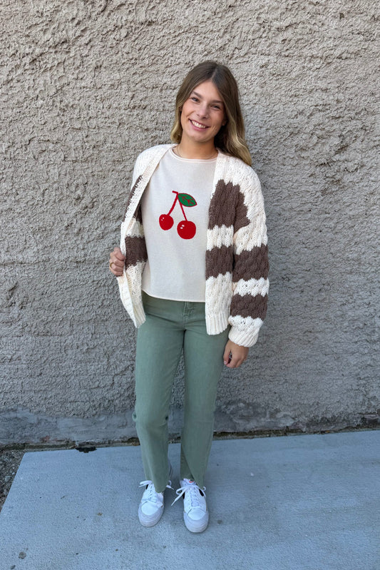 Person wearing a sweater with cherry design and a striped cardigan against a concrete wall.