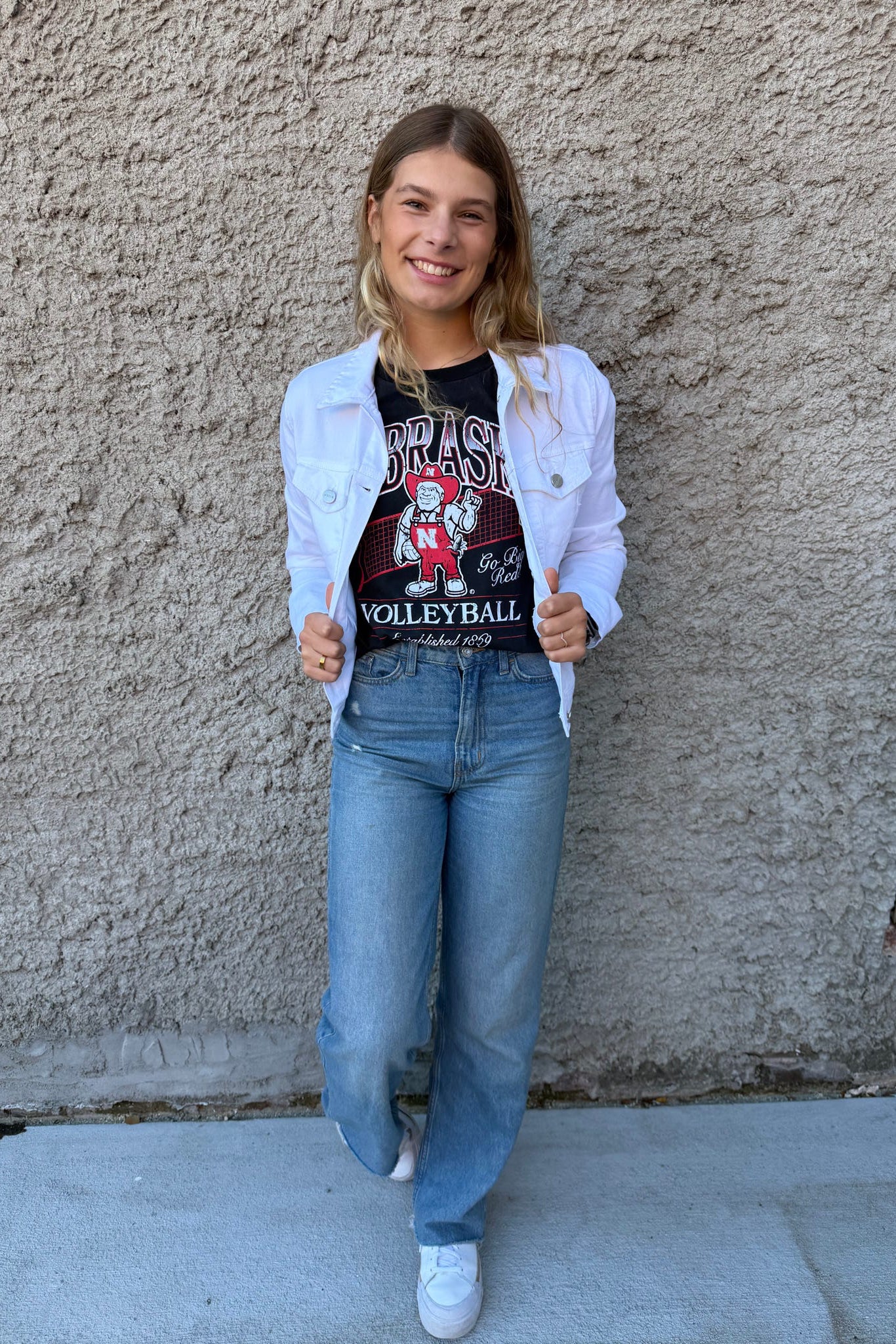 Person wearing a white jacket, black t-shirt with graphics, and blue jeans against a textured wall.