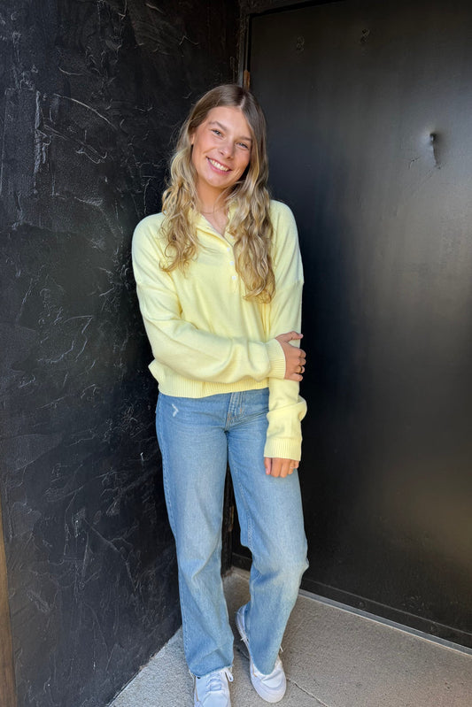 Woman wearing a yellow sweatshirt and blue jeans standing against a dark wall.