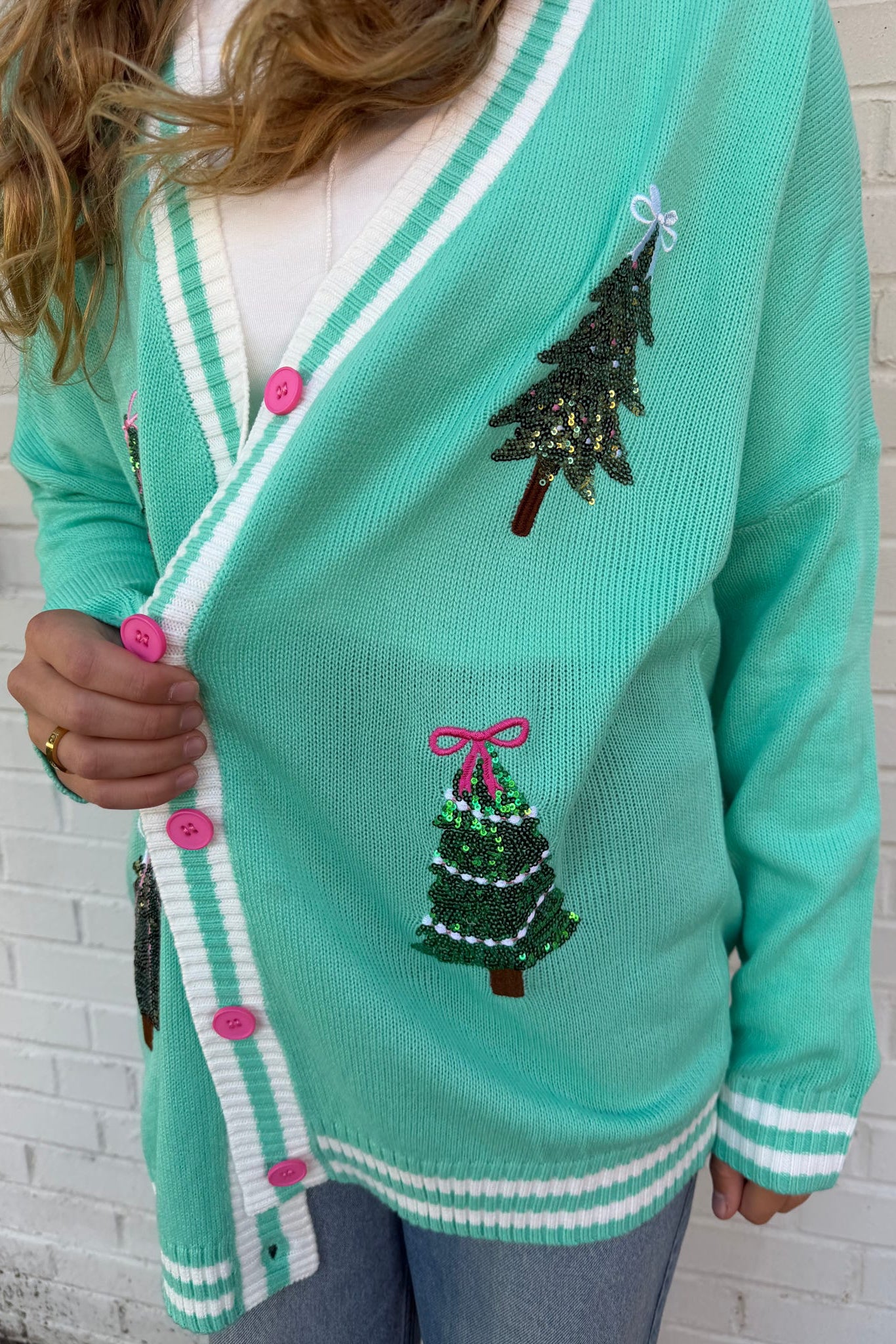 Teal cardigan with Christmas tree designs and buttons against a white brick wall.