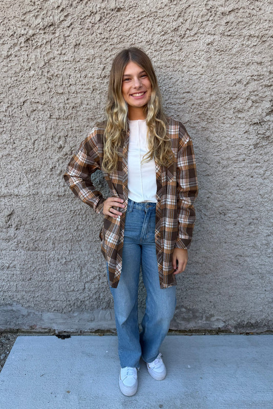 Person wearing a plaid shirt, white top, and jeans standing against a concrete wall.