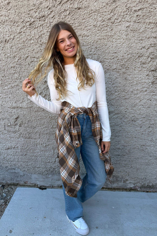Woman wearing a white long-sleeve top, blue jeans, and a plaid shirt over her shoulders against a beige wall.